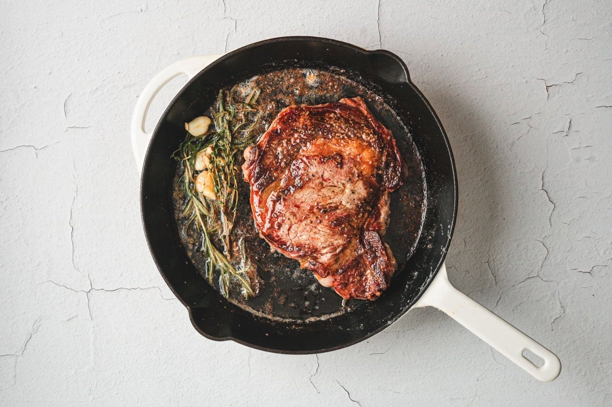 Pan seared ribeye steak in a cast iron pan with garlic, herbs, and butter.
