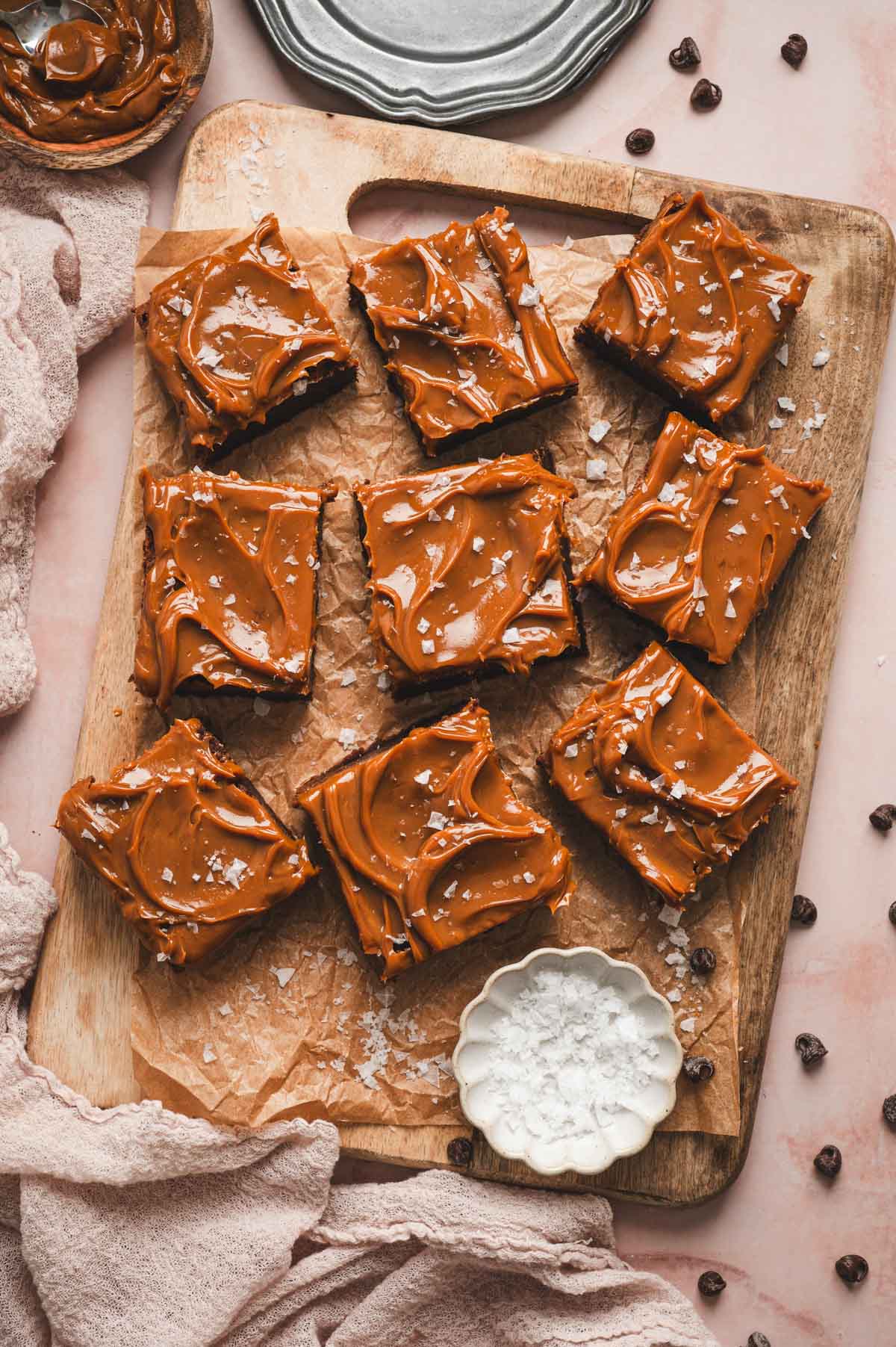 Wooden board with parchment paper topped with salted caramel brownies and white dish of flaky sea salt.