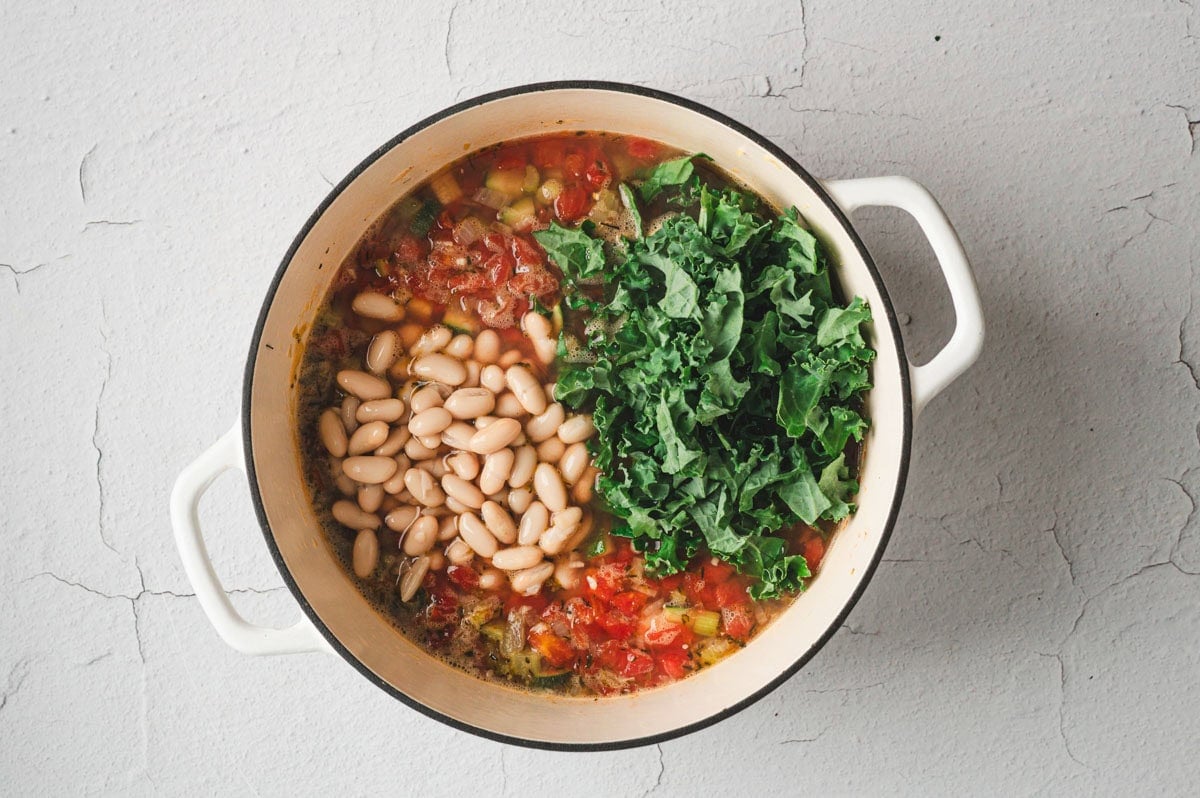 Beans and Kale added to soup in a white pot.