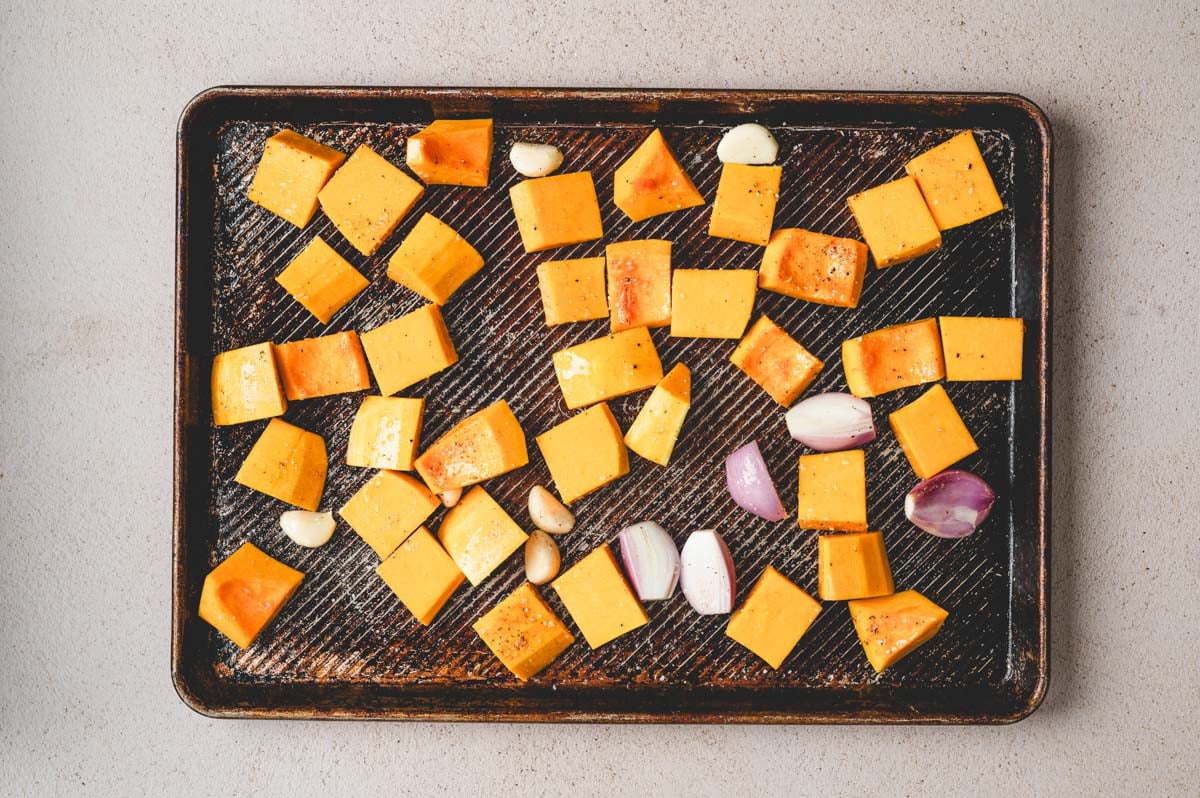 Butternut squash pieces, garlic cloves, and halved shallots on a sheet pan.