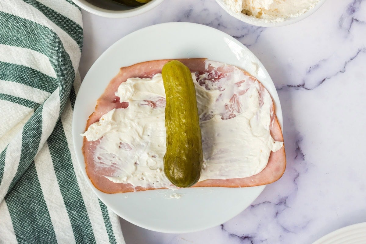 A ham slice on a plate topped with cream cheese spread and a whole pickle.