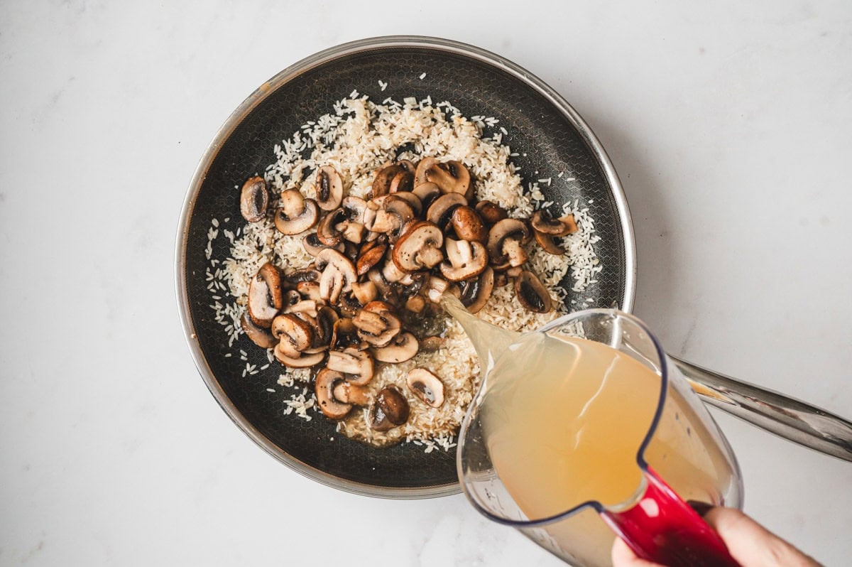 Rice with mushrooms in a skillet with broth being poured on top.
