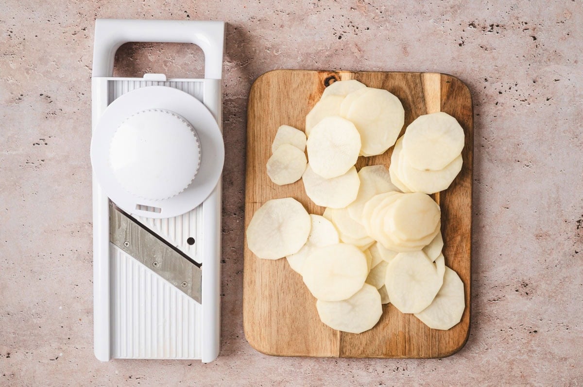 Potatoes sliced thinly on a wooden cutting board next to a white mandolin.