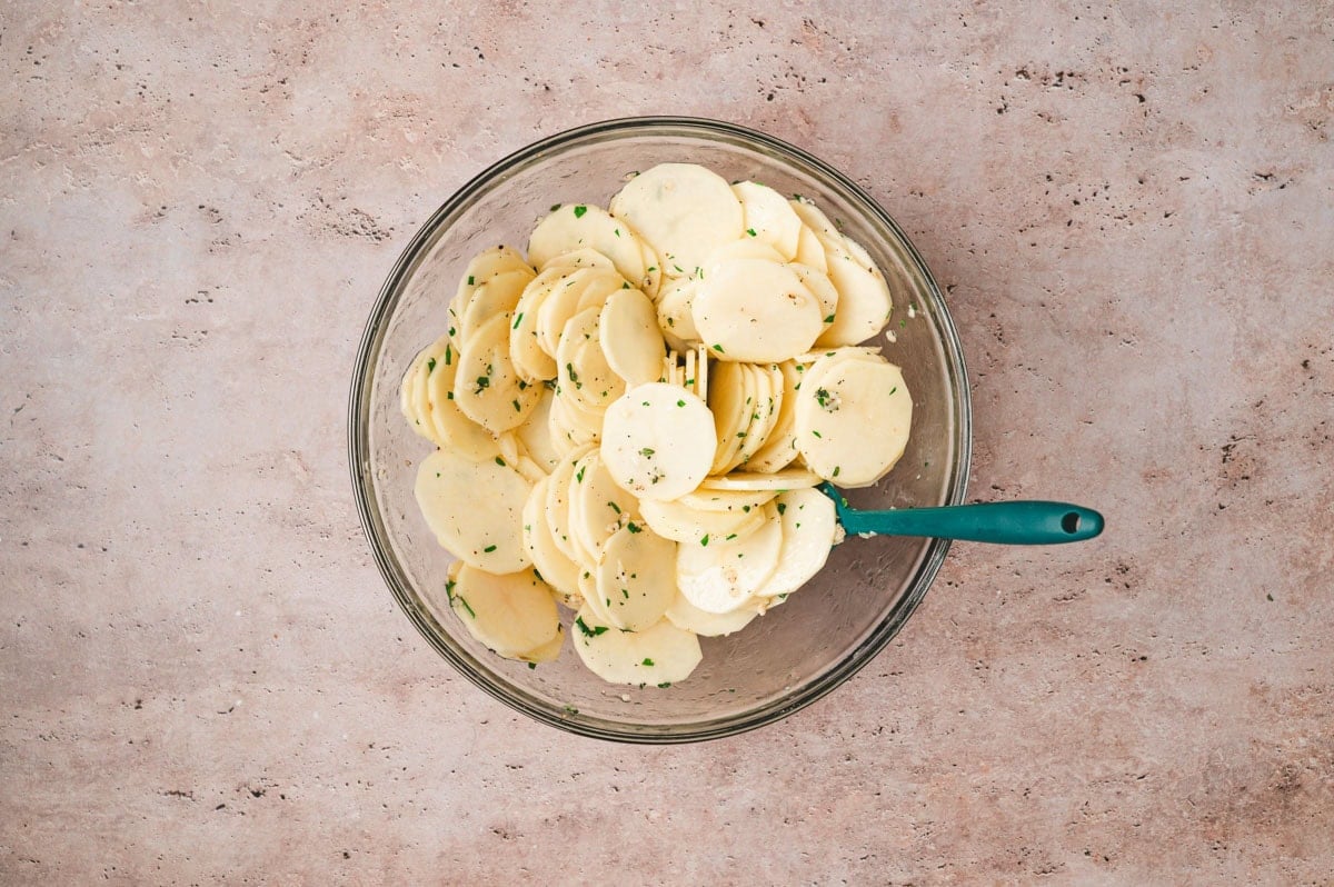 Thinly sliced potatoes tossed in butter, garlic and herbs in a glass bowl.