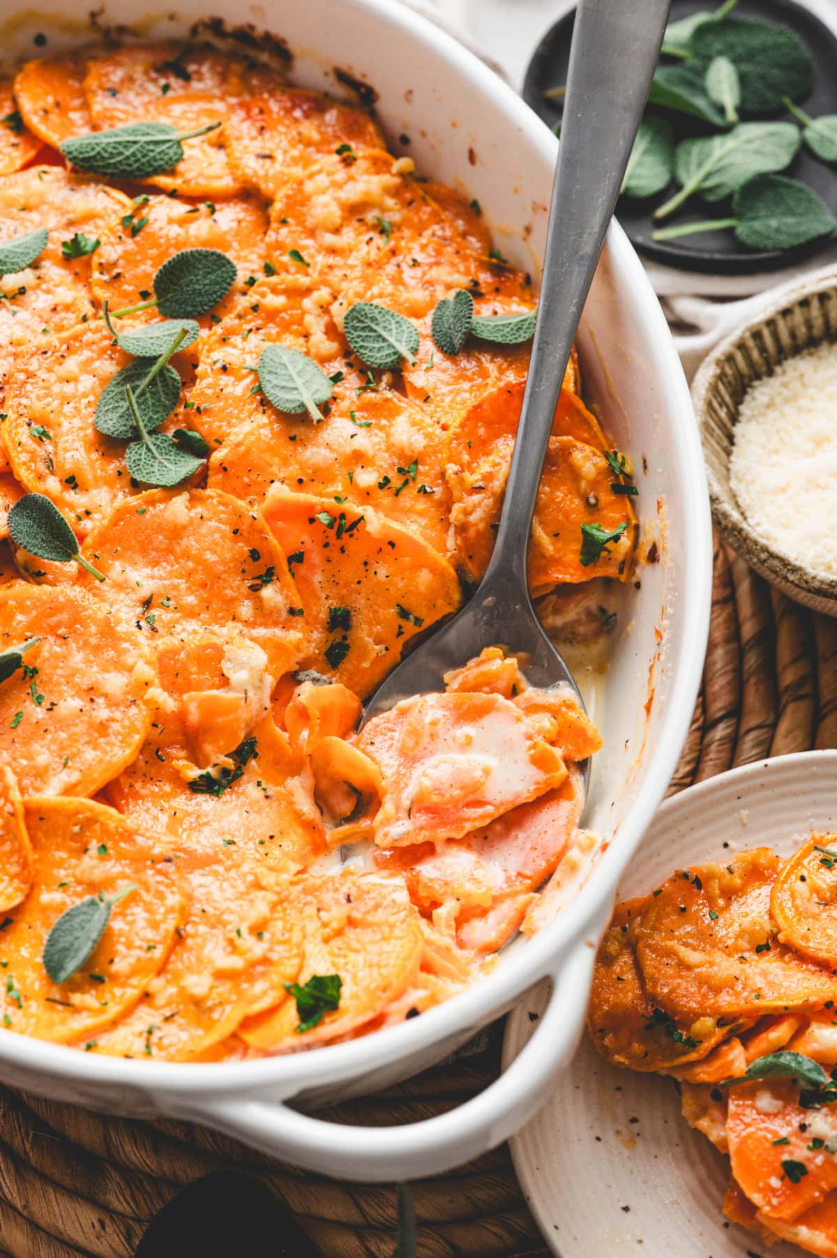 Sweet potato gratin in a white casserole dish with a serving spoon, garnished with sage leaves.
