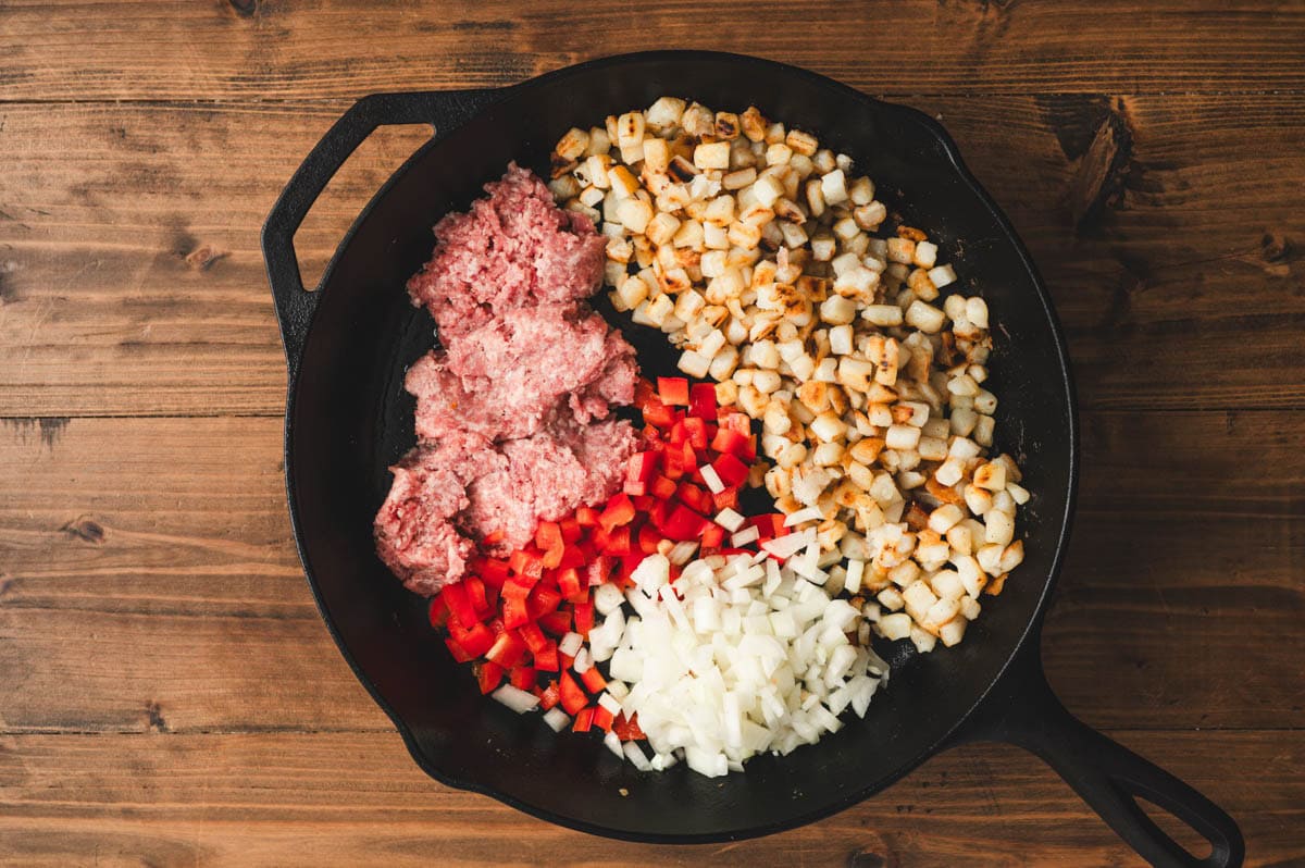 Cast iron skillet with cooked diced potatoes on one side, with sausage, diced peppers and onions on the other side.