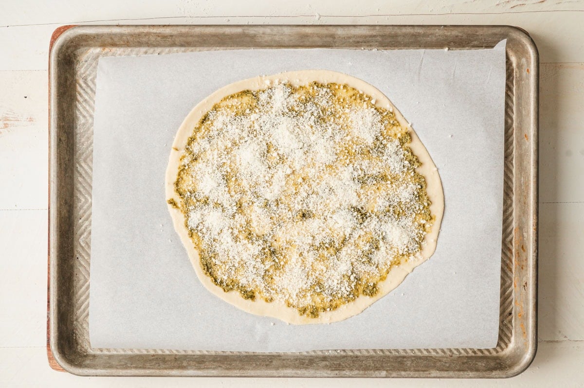 Flattened star bread dough with pesto and parmesan on top, all on a parchment paper lined sheet pan.