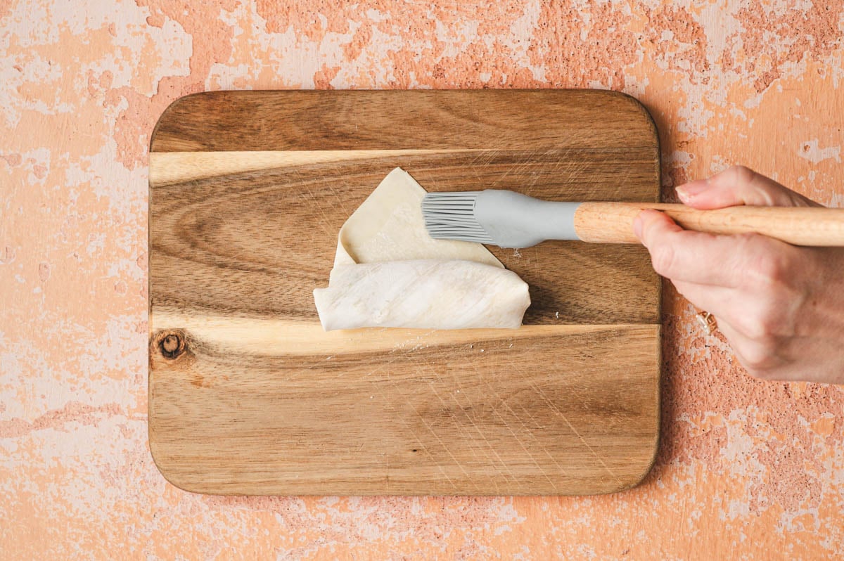 Uncooked egg roll being brushed with water while being rolled up.