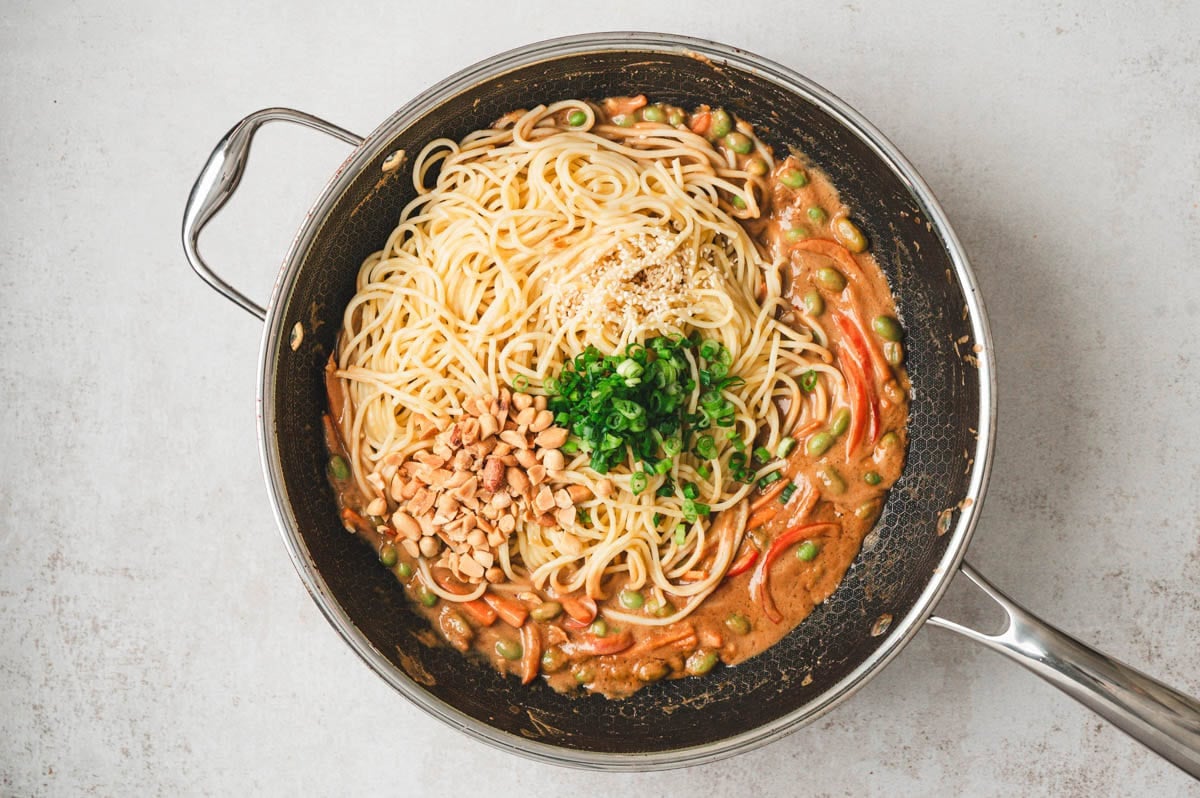 Cooked noodles, peanut sauce, veggies, chopped peanuts, sliced green onions, in a saute pan.