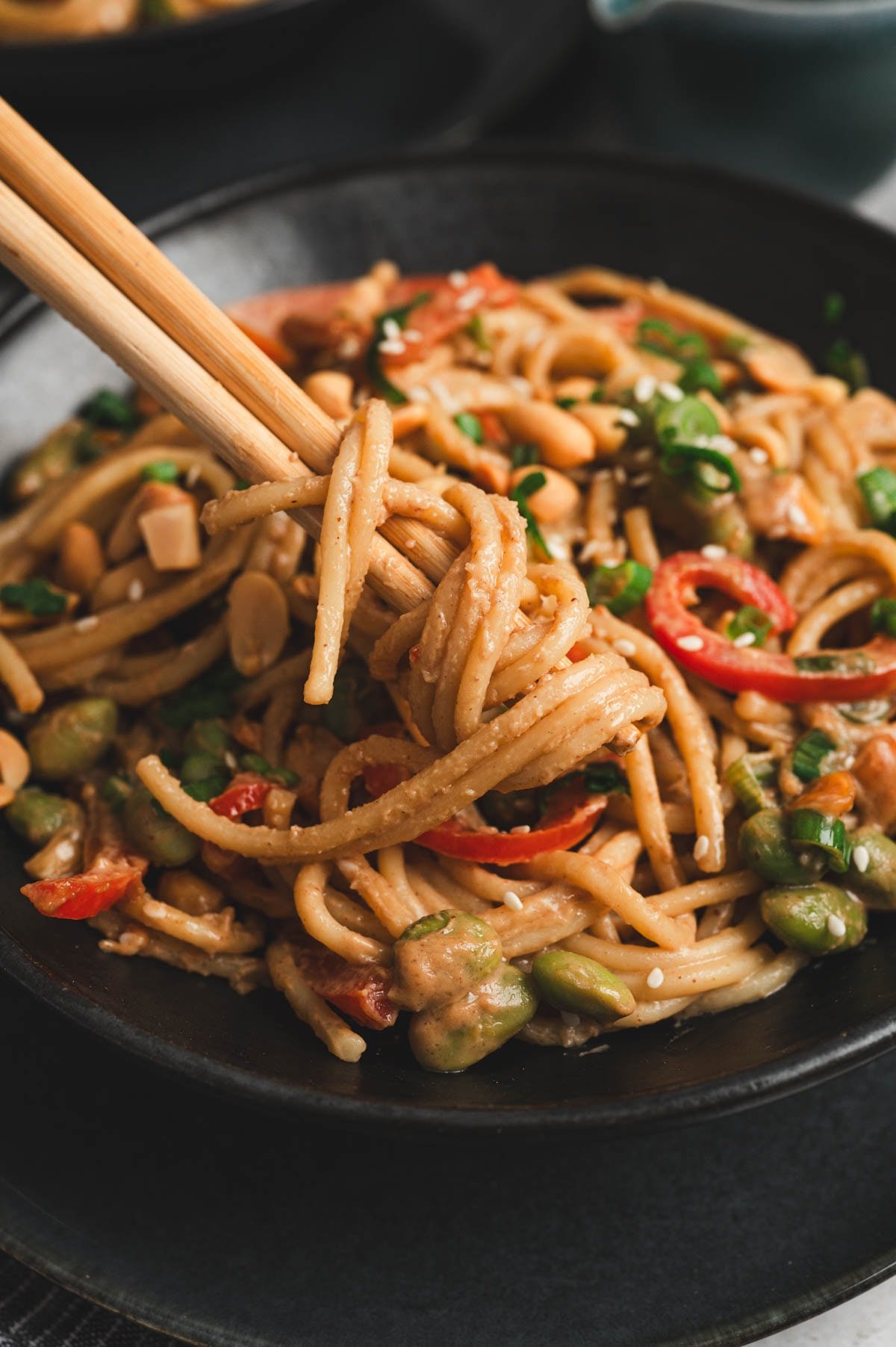 Noodles and veggies with peanut sauce in a dark bowl with wooden chopsticks.