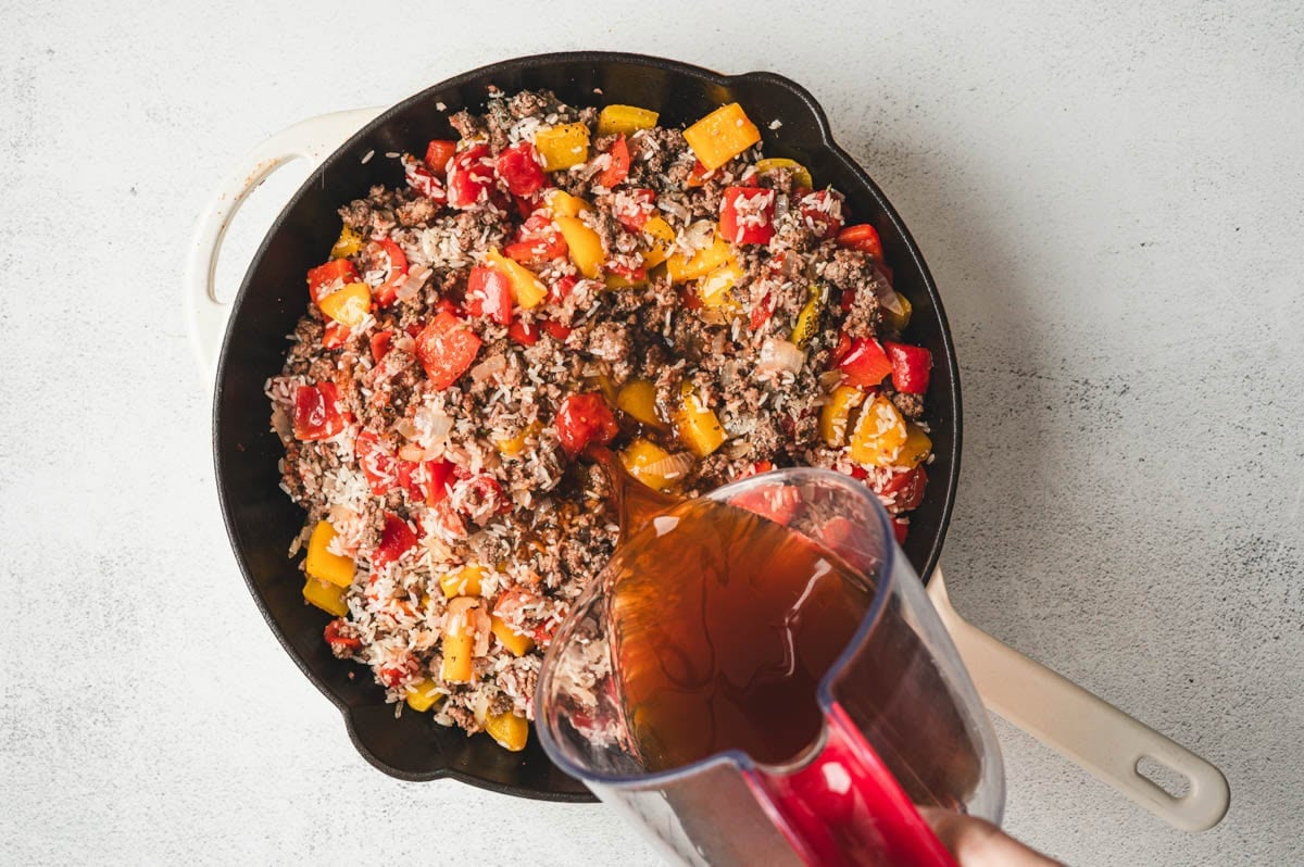 Skillet with ground beef, bell peppers, and uncooked rice. Beef broth being poured in.