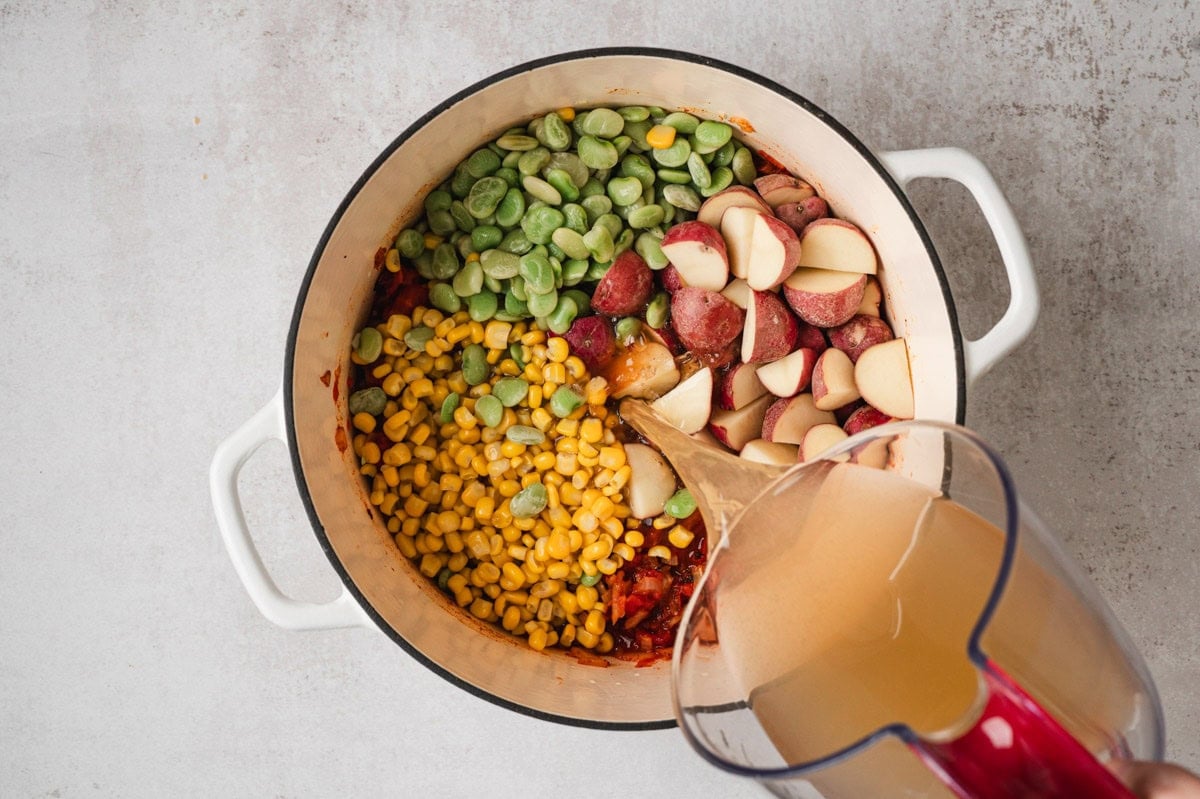 Ingredients for brunswick stew in a white pot with chicken broth being poured in.
