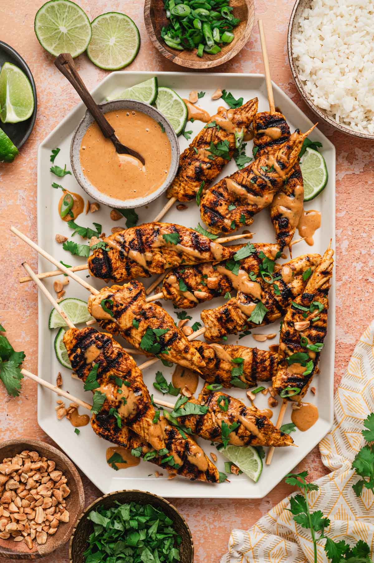 Chicken satay on wooden skewers drizzled with peanut sauce and garnished with cilantro and lime wedges.