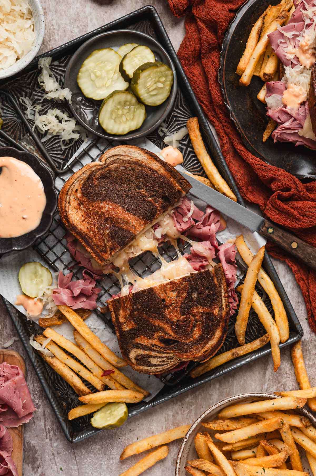 Reuben sandwich cut in half on a tray with fries, pickle slices and a knife.