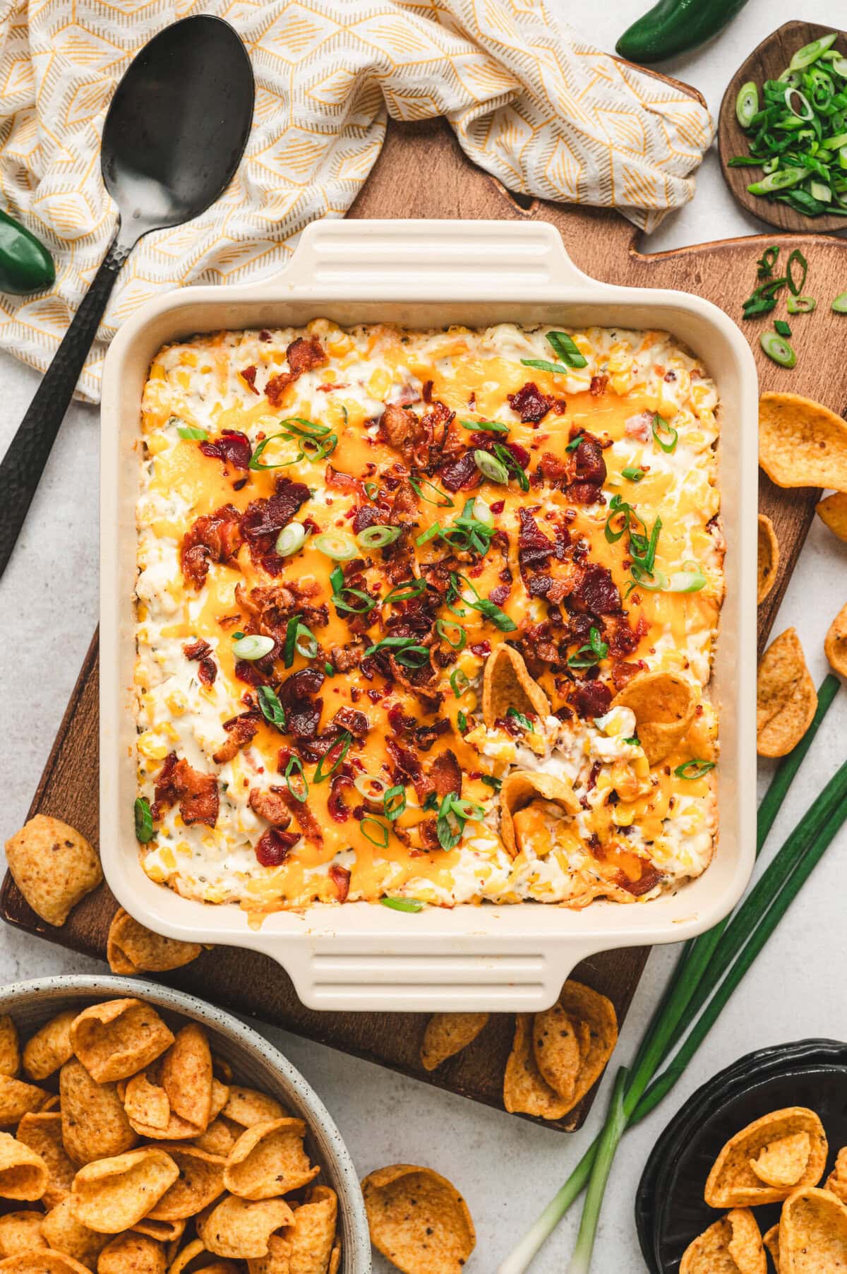 Square casserole dish with baked crack corn dip, garnished with green onions, with scoops tortilla chips