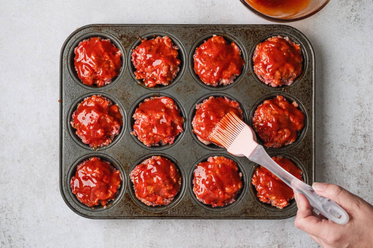 Raw mini meatloaves in a muffin tin with ketchup glaze being brushed on.
