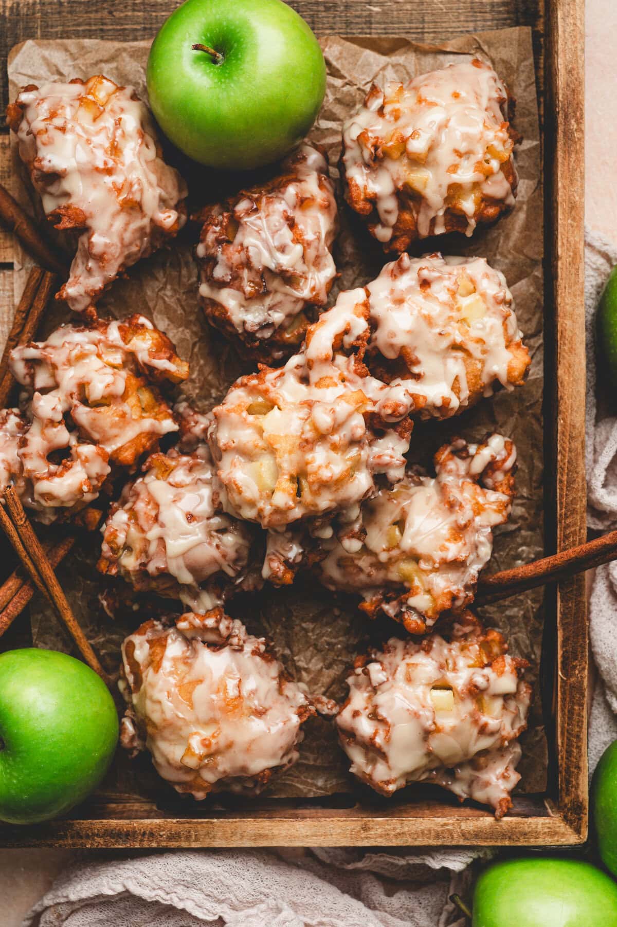 Apple fritters with glaze stacked on parchment paper next to granny smith apple and cinnamon sticks