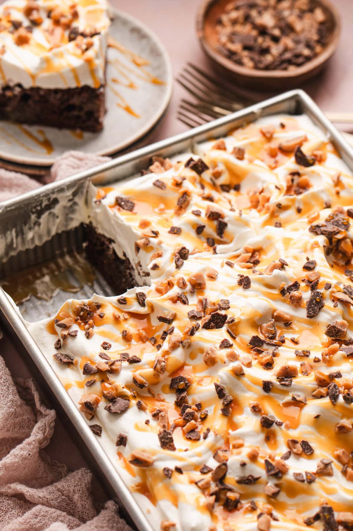 Angled shot of chocolate poke cake with whipped topping and caramel and toffee bits on top with a slice removed.