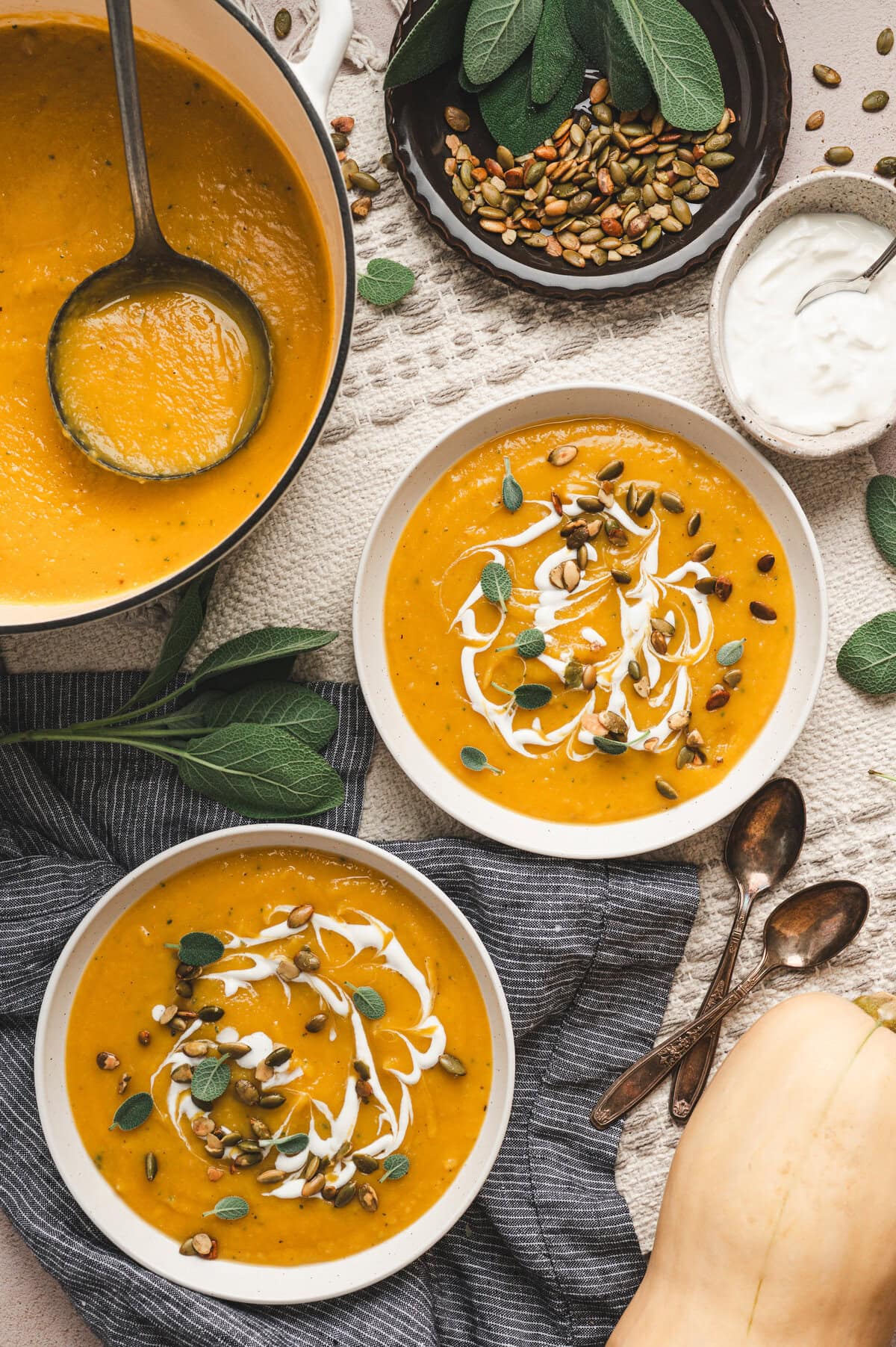 Two bowls of butternut squash soup topped with fresh sage, sour cream, and toasted pumpkin seeds