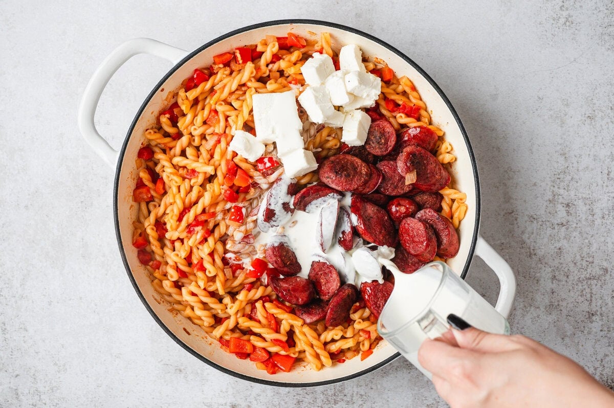 Cooked pasta in a pan with cream cheese, heavy cream, and smoked sausage in a white pan