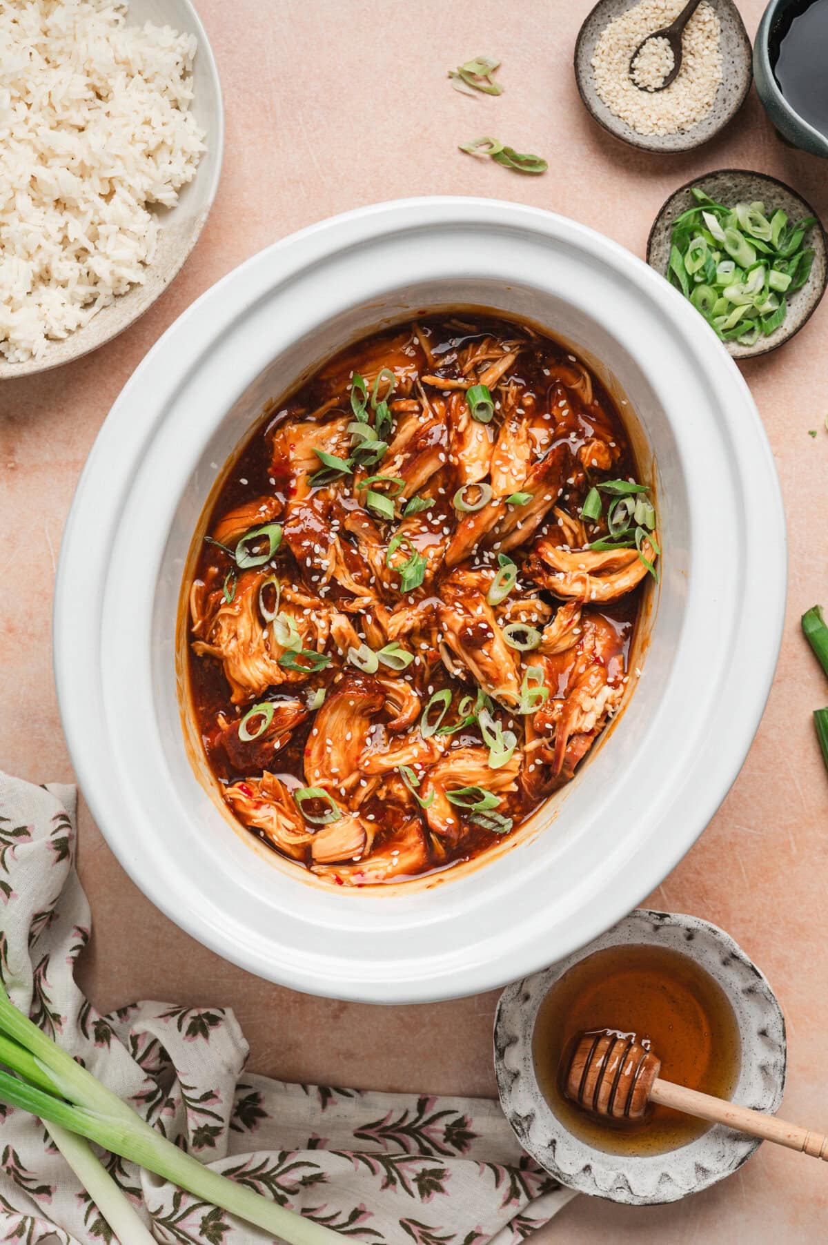 Cooked honey garlic chicken in a white slow cooker insert, garnished with green onions and sesame seeds